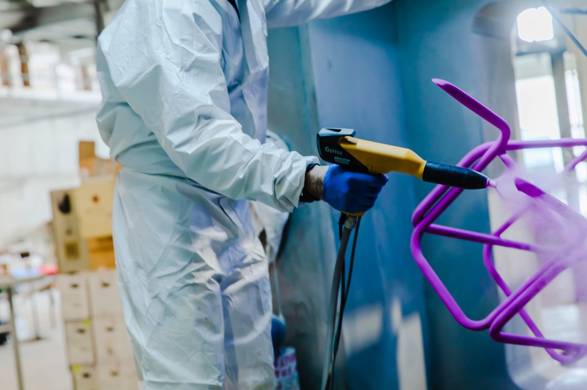 Detail of industrial worker using spray gun to apply powder coating on metal structure inside factory booth.